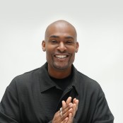 Paul C Brunson smiling towards the camera in front of a white backdrop, wearing a black tshirt and jacket